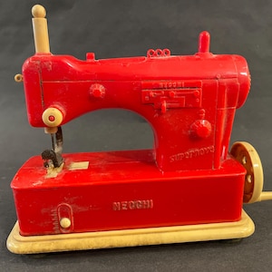 May include: A red toy sewing machine with a white base. The machine has a white handle, a black needle, and the words "SUPER TOY" and "NEcchi" printed on it.