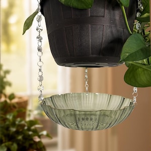 May include: A dark brown plastic hanging planter with a green leafy plant, suspended by clear crystal-like beaded chains that hold a scalloped, translucent sage green glass bowl underneath to catch water or display decorative items.