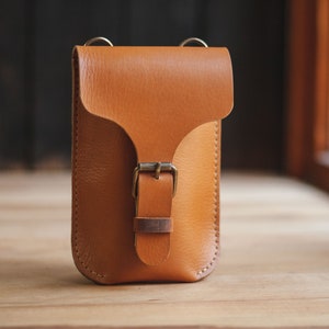 Op de afbeelding: Een klein, bruin leren schoudertasje met een gespsluiting. De tas heeft een verstelbare schouderband voor een comfortabele pasvorm.