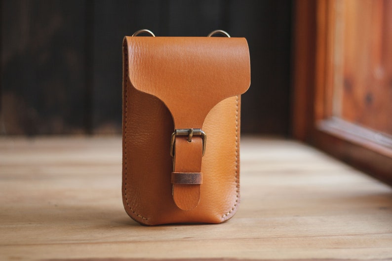 Op de afbeelding: Een klein, bruin leren schoudertasje met een gespsluiting. De tas heeft een verstelbare schouderband voor een comfortabele pasvorm.