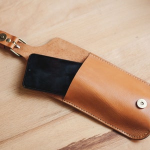 Op de afbeelding: Een bruine leren telefoonhoes met een band en een metalen knoop. Een zwarte smartphone zit in de hoes.