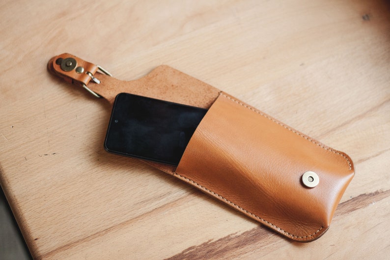 Op de afbeelding: Een bruine leren telefoonhoes met een band en een metalen knoop. Een zwarte smartphone zit in de hoes.