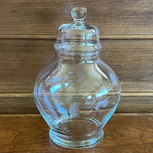 May include: A clear glass apothecary jar with a lid and a round knob. The jar has a bulbous body and a flared base. Delicate floral etchings adorn the glass. The jar is displayed on a wooden surface.