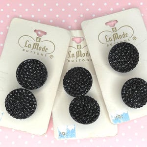 May include: Several black, textured buttons on white cards with the "La Mode Buttons" logo. The buttons have a raised, dotted surface. The cards are on a pink background with white polka dots. The price tag on the card reads "49¢".