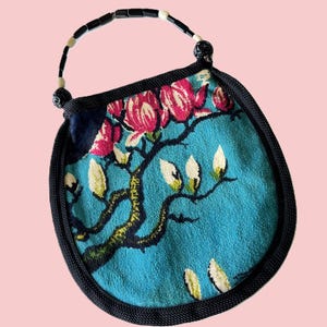 May include: A small, round handbag with a beaded handle. The bag features a teal background with a floral design of pink flowers and white buds on a dark branch. The bag is trimmed with black fabric.