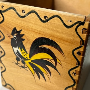 Peut inclure: Boîte en bois avec un motif de coq peint à la main. Le coq est noir, jaune et blanc, avec une queue détaillée. La boîte a une bordure décorative noire ondulée avec de petits points. La boîte est en bois de couleur claire.