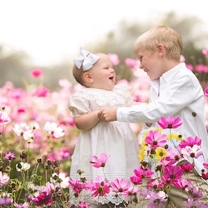 Peut inclure: Une jeune fille en robe blanche avec un nœud blanc dans les cheveux rit en regardant un jeune garçon en chemise blanche. Ils sont debout dans un champ de fleurs roses et blanches.