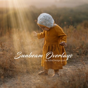 May include: A young child wearing a yellow dress and a floral print bonnet stands in a field of tall grass. The sun shines through the grass, creating a warm glow. The text "Sunbeam Overlays by AsheDesign.com" is visible in the lower center of the image.