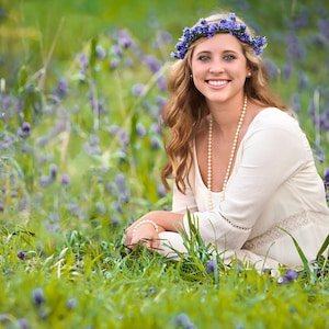 May include: A young woman with long brown hair wearing a white dress and a flower crown sits in a field of purple wildflowers.