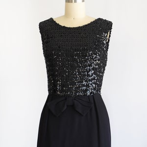 Side front view of a vintage late 50s early 60s black dress that has a black sequin bodice, a bow at the waist, and a pencil skirt. The dress is modeled on a dress form