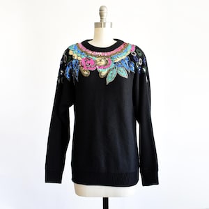 May include: Black crew neck sweater with long sleeves. The neckline and shoulders are adorned with a floral sequin design in pink, blue, and green. The garment is displayed on a mannequin against a white backdrop.