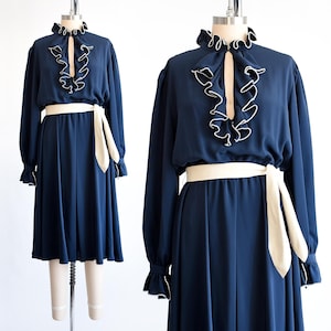 Puede incluir: Un vestido azul marino con cuello de volantes blanco y cinturón blanco. El vestido tiene mangas largas y una falda fluida.