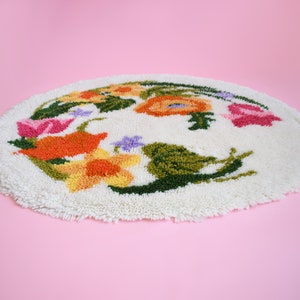 A view from the side of a vintage round floral latch hook rug that has cream white shaggy acrylic with a colorful floral motif in pinks, orange, yellow, purple and greens. The rug and the rug is on a pink background.