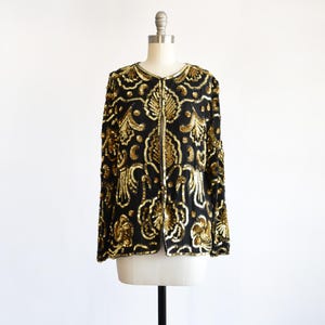 May include: Black jacket with gold sequin embellishments in an ornate floral pattern. The jacket is displayed on a mannequin. The jacket has long sleeves and a round neckline. The sequins create a shimmering effect.