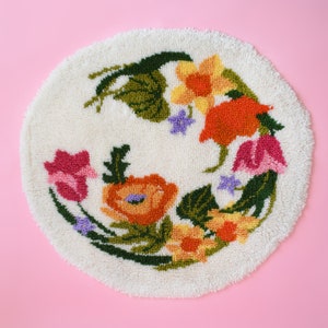 A vintage round floral latch hook rug that has cream white shaggy acrylic with a colorful floral motif in pinks, orange, yellow, purple and greens. The rug is on a pink background.