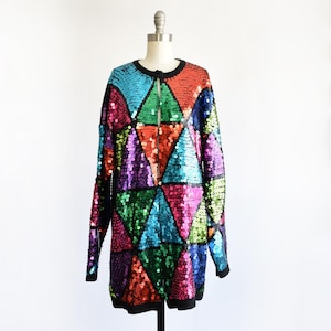 May include: A long-sleeved, sequined jacket with a geometric pattern of colourful triangles. The jacket has a black collar and trim, with a button closure at the neckline. The sequins are in shades of blue, green, orange, pink, and purple.