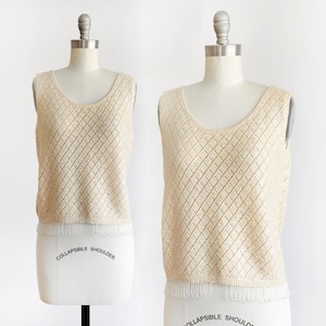 May include: A sleeveless, cream-colored top with a scoop neck and a diamond-patterned knit design. The bottom edge features a fringe detail. The top is displayed on a mannequin with the words "COLLAPSIBLE SHOULDER" printed on the base.