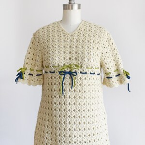 Side front view of a vintage 70s crochet mini dress with a green and blue woven stripe on the front that ties into a bow, along with matching stripes and bows on the sleeves. The dress is on a dress form.