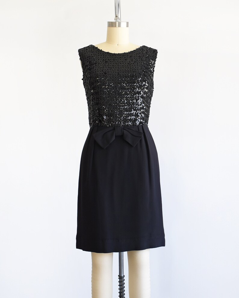 A vintage late 50s early 60s black dress that has a black sequin bodice, a bow at the waist, and a pencil skirt. The dress is modeled on a dress form