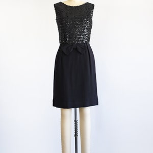 A vintage late 50s early 60s black dress that has a black sequin bodice, a bow at the waist, and a pencil skirt. The dress is modeled on a dress form