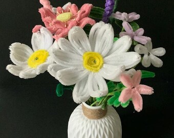 May include: A white ceramic vase with a textured surface holds a bouquet of pipe cleaner flowers. The flowers are white, pink, and purple.