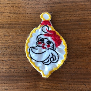May include: Embroidered Christmas ornament featuring a cartoon Santa Claus face. The ornament has a white background with a yellow scalloped border. Santa wears a red hat and has a black outline. A festive holiday decoration.