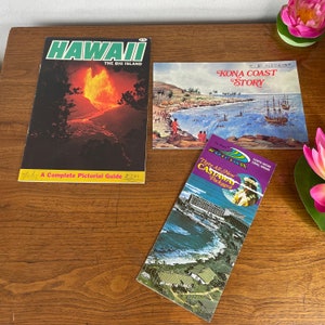 May include: Three vintage travel brochures about Hawaii. The first brochure features a red, orange, and yellow volcanic eruption with the text "Hawaii, The Big Island". The second brochure features a blue ocean scene with a sailboat and the text "Kona Coast Story". The third brochure features a blue ocean scene with a hotel and the text "Three All New Castaway Packages".