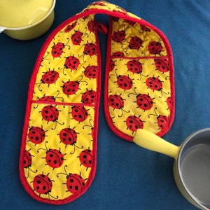 May include: A pair of yellow oven mitts with a red trim and a ladybug pattern. The mitts are connected by a yellow fabric loop.