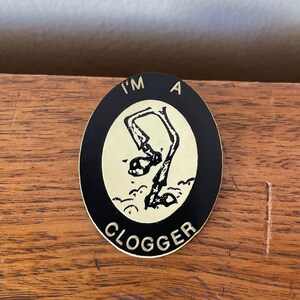 May include: An oval-shaped pin with a black border and a cream-colored center. The pin features a black illustration of a person diving into water, with the words "I'M A" at the top and "CLOGGER" at the bottom.