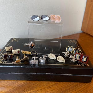 May include: A selection of vintage cufflinks in diverse styles. Features silver, gold, and enamel designs, some with geometric patterns, others with intricate details. Displayed on a black box with a decorative lid.