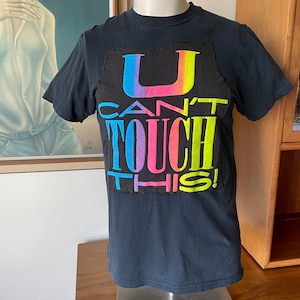 Puede incluir: Camiseta negra con la frase "U CAN'T TOUCH THIS!" en un diseño multicolor. El texto presenta un degradado de arcoíris. La camiseta tiene mangas cortas y cuello redondo. Un parche negro está cosido en la parte delantera.