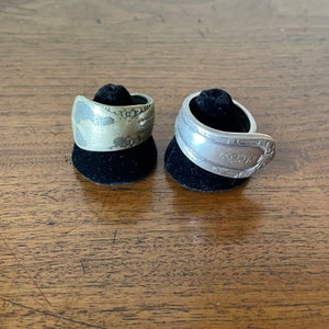 May include: Two spoon rings, one silver and one tarnished, rest on black velvet ring displays. The silver ring has a floral design, while the tarnished ring has a stamped pattern. Both rings are on a wooden surface.