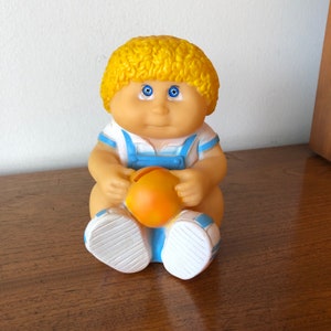 May include: A vintage Cabbage Patch Kid piggy bank with blonde hair, blue eyes, and a blue and white outfit. The piggy bank is holding a yellow ball.