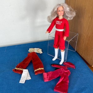 May include: A vintage Charlie's Angels doll with long blonde hair, wearing a red jumpsuit with the words "Charlie's Angels" on the front. The doll is holding a white purse and is wearing white boots. The doll is also wearing a white belt and a white headband. The doll is standing on a clear plastic stand.