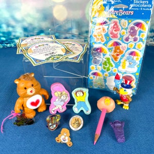 May include: A collection of Care Bears themed items including a brown bear figurine, a pencil with a pink eraser shaped like a bear face, a sticker sheet with Care Bears characters, and other small toys.