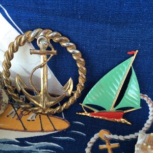 May include: Two gold-tone nautical themed brooches. One is a round anchor with a rope design. The other is a green sailboat with a red bottom.