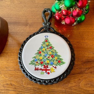 May include: A round, white ceramic trivet with a black metal frame and handle. The trivet features a colorful Christmas tree design with ornaments, gifts, and a toy train. The word "ACTION" is printed on the trivet.