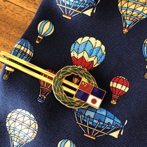 May include: A gold tie clip with a wreath and a blue, white, and red square with a Japanese flag in the lower right corner. The tie clip is on a navy blue tie with a pattern of hot air balloons.