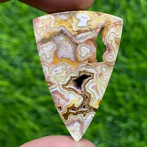 May include: A triangular shaped agate cabochon with a brown, white, and yellow banded pattern. The stone has a natural, rough edge and a smooth, polished surface.