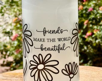 May include: A white glass can-shaped tumbler with a clear lid. The tumbler features a black floral design and the words "friends make the world beautiful" in a script font. The background is blurred.