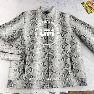 Op de afbeelding: Een leren jas met slangenpatroon in grijstinten en wit. De jas heeft een ritssluiting, twee voorzakken en de tekst "UMAROCS LEATHER" in een cirkelvormig ontwerp op de borst. De tekst "WhatsApp +44 7544 336431" is ook zichtbaar.