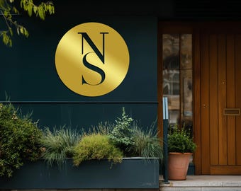 May include: A gold circular sign with the black letters "NS" on a dark teal wall. Below the sign is a rectangular planter with various green plants. A wooden door and a terracotta pot with a plant are in the background.