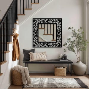 May include: A black, square mirror with a decorative, openwork frame hangs on a white wall. The mirror reflects a staircase. Below, a bench holds pillows, a basket, and a rug. A large vase with greenery sits to the right.