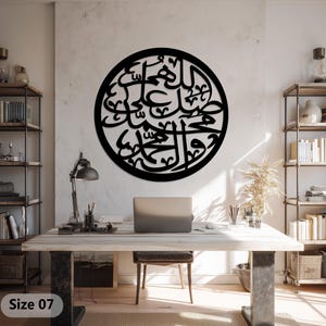 May include: A large, black, circular wall art piece featuring Arabic calligraphy. The artwork is mounted on a light-colored wall above a wooden desk. The desk has a laptop, lamp, and office supplies. Bookshelves are on either side.