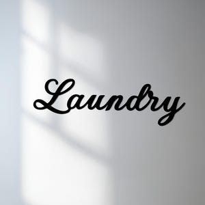 Può includere: Cartello nero "Laundry" in corsivo. La parola è ritagliata da un materiale solido e montata su un muro bianco. L'insegna è in un carattere script e proietta un'ombra.