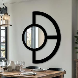 May include: A modern, geometric wall mirror with a black frame. The design features a circular mirror element within a larger, abstract shape. The mirror is mounted on a white wall, reflecting the room's interior. The overall aesthetic is contemporary and minimalist.