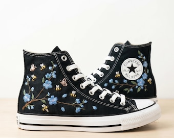 May include: Black high-top sneakers with white laces and soles. The sneakers feature embroidered blue flowers, bees, and butterflies. The Converse All Star logo is visible on the side.