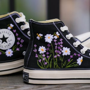 Puede incluir: Zapatillas de lona negras de caña alta con cordones blancos y suela de goma blanca. Las zapatillas están bordadas con margaritas blancas, lavanda morada y abejas amarillas.