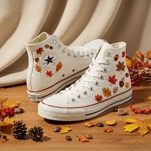 May include: Pair of white high-top sneakers with embroidered autumn leaves and acorns. The sneakers have a red stripe on the sole and the Converse All Star logo. The shoes are on a wooden surface with fall foliage.