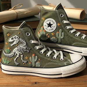 May include: Olive green high-top sneakers with white laces and soles. The sneakers feature embroidered designs of a dinosaur skeleton, cacti, and star accents. A Converse All Star patch is on the side.